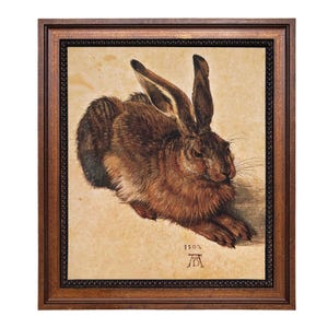 May include: A framed print of a brown hare, a detailed artwork with long ears and fur. The artwork is set against a light background, framed in a dark wood frame. The year 1502 and an artist's mark are visible.