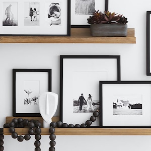 May include: Two horizontal wooden wall shelves holding black picture frames with black and white photographs, a white abstract face sculpture, a string of dark wooden beads, and a rectangular gray planter containing a dark succulent plant.