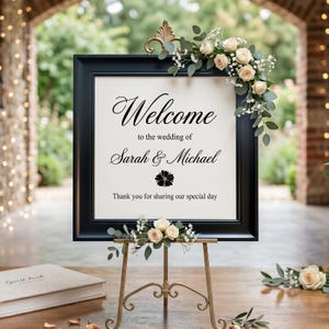 May include: A black framed wedding sign with the text "Welcome to the wedding of Sarah & Michael". The sign is decorated with cream-colored roses and greenery, and sits on a gold easel.