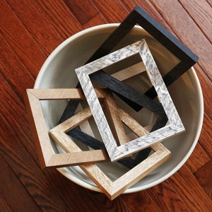 May include: A collection of five picture frames in a white bowl. The frames are made of wood and have a variety of finishes, including black, brown, and gray. The frames are arranged in a random order, with some overlapping.