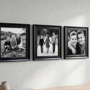 May include: Three black framed photographs of children hanging on a white wall. The photos are black and white and show children in various poses. The frames are square with a simple, elegant design. The photos are hung above a wooden surface.