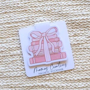 May include: A magnetic bookmark with a design of a stack of pink books tied with a bow. The bookmark has a white background and the text "Mamas Creations MAGNETIC BOOKMARK". The design is on a white card with a clip.