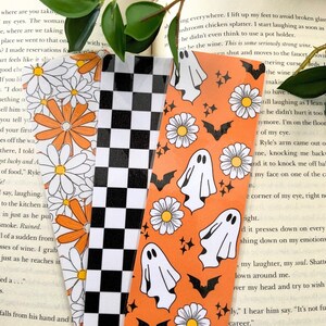 May include: Three Halloween themed bookmarks. One bookmark features a black and white chequered pattern. The other two bookmarks feature a bright orange background with white daisies, white ghosts, and black bats.