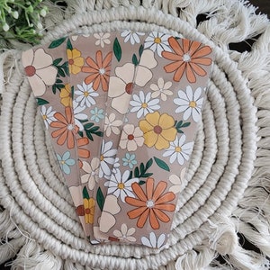 May include: A stack of floral-patterned bookmarks with a retro daisy design in orange, yellow, white, and green on a tan background. The bookmarks are on a cream macrame coaster, with greenery in the background.
