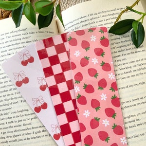 May include: Three bookmark designs featuring a red and white checkered pattern, a pink background with red strawberries and white flowers, and a pink background with red strawberries and white flowers. The bookmarks are made of clear plastic.