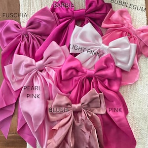 May include: An assortment of satin bows in shades of pink, such as fuchsia, Barbie, bubblegum, pearl pink, blush, light pink, and pink. Each bow is labelled with its colour name.