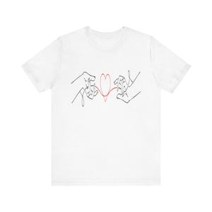 Puede incluir: Camiseta blanca con un diseño minimalista de arte lineal. El diseño representa dos manos conectadas por un corazón rojo. Las manos están dibujadas con líneas negras, creando un gráfico simple pero impactante.