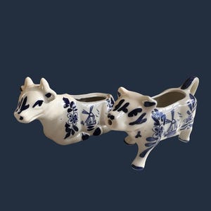 Può includere: Due lattiere in ceramica color crema a forma di mucca, decorate con motivi floreali e illustrazioni di mulini a vento dipinti in blu. Una mucca è in posizione sdraiata, mentre l'altra è in piedi con un'apertura sulla bocca per versare.