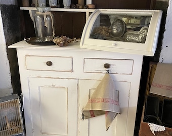 Op de afbeelding: Wit geschilderd kast met twee laden en twee deuren. De kast heeft een verweerde afwerking en is versierd met een linnen handdoek. De kast staat op een houten vloer.