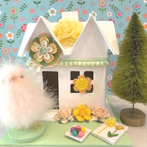 May include: A white decorative house with a green base, adorned with paper flowers and a small green bottle brush tree. A fluffy chick and a small gift box are also present. The background features a floral pattern.