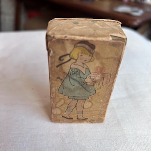 May include: A rectangular cardboard box with a vintage-style illustration of a child with blonde hair wearing a dark cap and a light blue outfit. The child is holding a gift box with a bow. The box has a tan background with small colorful dots.