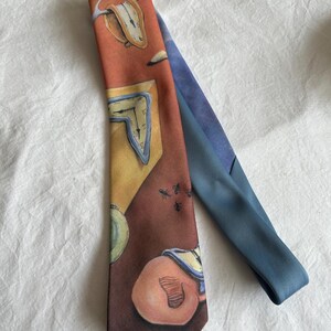 May include: A necktie featuring a surrealist design with melting clocks, ants, and other abstract elements. The tie has a color palette of orange, brown, yellow, and blue. The tie is made of a smooth, likely silk or satin material.