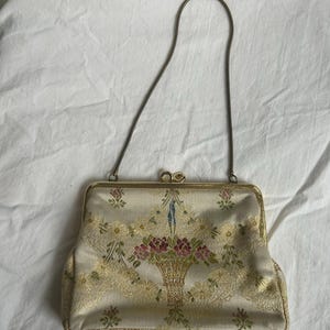 Pode incluir: Uma bolsa vintage creme com um design de cesta floral. A bolsa apresenta uma estrutura de metal dourado e uma alça delicada. O padrão floral inclui detalhes em rosa, verde e amarelo, criando um visual clássico e elegante.