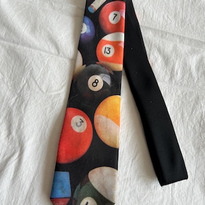 May include: A black necktie featuring a graphic print of various colorful billiard balls, including the number 8 ball, the number 13 ball, the number 14 ball, and a pool cue. The tie has a solid black back and a pointed tip at the bottom end.