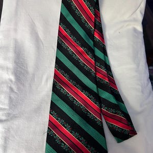 May include: A necktie featuring a diagonal striped pattern in black, bright green, and red. The black sections have a subtle herringbone texture, while the green sections are adorned with a repeating pattern of small green holly leaves and red berries.