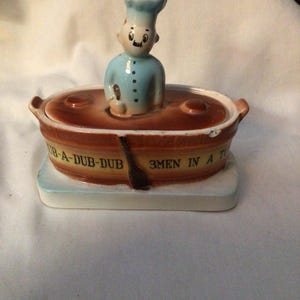 May include: A vintage ceramic figurine featuring a chef in a blue uniform emerging from a brown oval dish. The dish has the text "RUB-A-DUB-DUB 3MEN IN A TUB". The figurine is on a rectangular base.