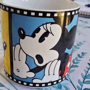 May include: A white ceramic mug with a yellow, blue, and red design featuring Mickey Mouse and Minnie Mouse. The design includes a film strip border.