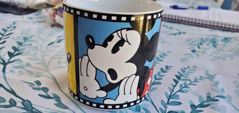 May include: A white ceramic mug with a yellow, blue, and red design featuring Mickey Mouse and Minnie Mouse. The design includes a film strip border.