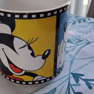 May include: A white ceramic mug with a black and yellow design featuring Minnie Mouse. The design includes a film strip border and Minnie Mouse's face with a red smile.