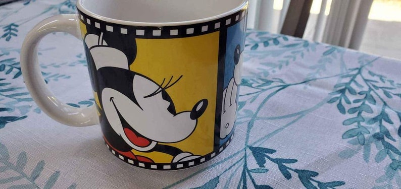 May include: A white ceramic mug with a black and yellow design featuring Minnie Mouse. The design includes a film strip border and Minnie Mouse's face with a red smile.