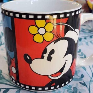 May include: A white ceramic mug with a black and red design featuring Mickey Mouse and Minnie Mouse. The design includes a yellow flower and a black and white film strip border.
