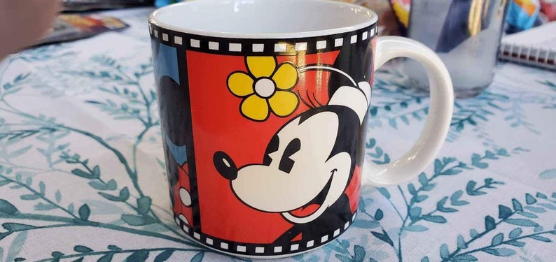 May include: A white ceramic mug with a black and red design featuring Mickey Mouse and Minnie Mouse. The design includes a yellow flower and a black and white film strip border.