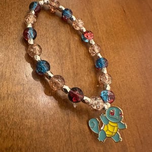 May include: A beaded bracelet featuring a Squirtle charm. The bracelet has a mix of clear, blue, red, and brown beads. The Squirtle charm is blue, yellow, and gold. The bracelet is on a wooden surface.