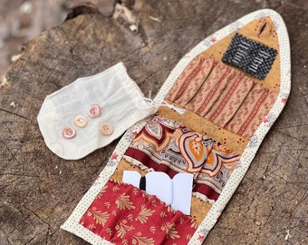 May include: A fabric needle case with multiple pockets and compartments. The case is made of patchwork fabric in shades of red, brown, and gold. The case is open and shows a small white card inside. A small white bag with buttons is next to the case.