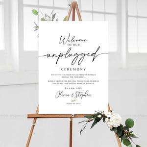 May include: A white sign on a wooden easel with the words "Welcome to our unplugged ceremony." The sign also includes a request to turn off phones and cameras. The names Olivia & Stephen and the date August 18, 2025 are also on the sign.