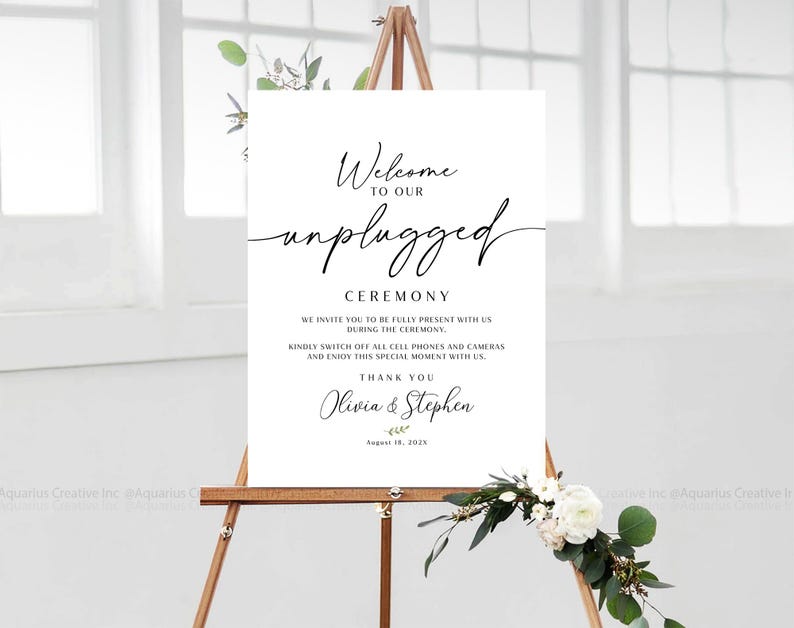 May include: A white sign on a wooden easel with the words "Welcome to our unplugged ceremony." The sign also includes a request to turn off phones and cameras. The names Olivia & Stephen and the date August 18, 2025 are also on the sign.
