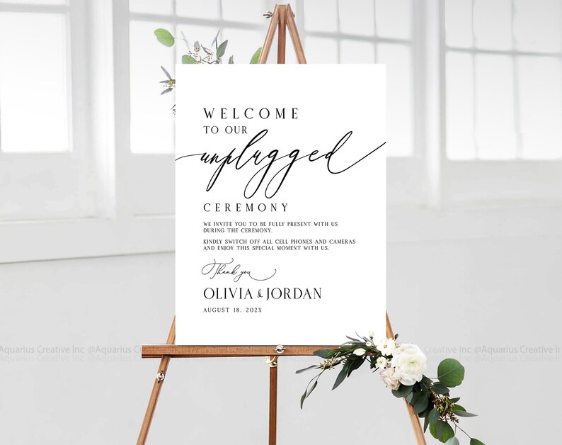 May include: A white sign on a wooden easel with the words "Welcome to our unplugged ceremony." The sign also includes a request to turn off cell phones and cameras. The names Olivia & Jordan and the date August 18, 202X are also on the sign.