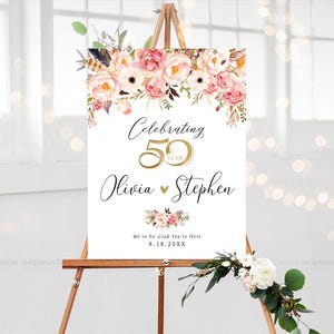 May include: A white sign on a wooden easel, adorned with pink and white floral arrangements. The sign displays "Celebrating 50 Year" with the names "Olivia & Stephen" and the date "8.18.20XX".