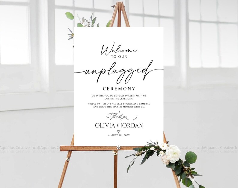 May include: A white sign with black text that reads 'Welcome to our unplugged ceremony. We invite you to be fully present with us during the ceremony. Kindly switch off all cell phones and cameras and enjoy this special moment with us. Thank you Olivia & Jordan ♡ August 18, 2023' The sign is propped up on an easel with greenery and white flowers in the foreground.