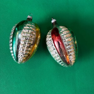 May include: Two vintage Christmas ornaments shaped like corn cobs. One is green and gold, the other is silver with a red stripe. Both have a textured surface and silver metal caps for hanging. The ornaments are displayed on a green background.