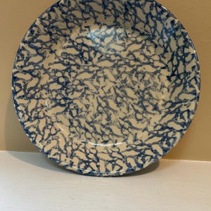 May include: A round ceramic bowl with a cream-colored base and a blue, intricate, web-like pattern. The bowl has a slightly raised rim and sits on a white surface. The bowl is likely for serving food.