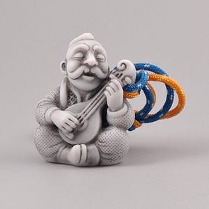 May include: A small, light gray figurine of a seated person with a mustache playing a stringed instrument. The figure has a textured surface and is attached to a loop of thick, braided blue and orange rope extending from the instrument's neck.