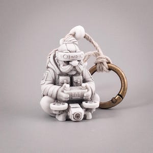 May include: A white, sculpted figurine keychain depicting a character with a mustache wearing a headset labeled MD, a tactical vest, and holding a remote control for a drone with two propellers. The figure is attached to a bronze-colored metal ring.