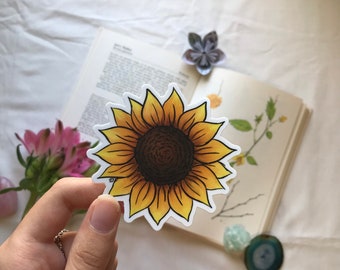May include: A yellow and brown sunflower sticker with a white background. The sticker is held in a hand.
