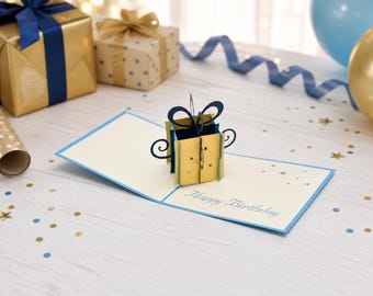 May include: A cream-colored pop-up greeting card featuring a three-dimensional yellow gift box with a dark blue bow. The card is open, displaying the text Happy Birthday in a script font, accented with small blue and gold star-shaped cutouts.