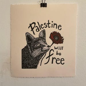 Peut inclure: Estampe en linogravure en noir et blanc représentant un renard avec une fleur rouge et l'inscription "Palestine will be free". L'estampe est sur papier crème et fixée par une pince.