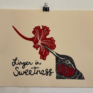 May include: A print showing a black hummingbird with a red throat and a red flower. The text "Linger in Sweetness" is written in black script below the flower and bird. The print is on cream-coloured paper.