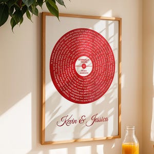 Peut inclure: Impression encadrée représentant un vinyle rouge avec du texte blanc. Le cadre en bois affiche les noms "Kevin & Jessica" en cursive. L'impression est accrochée au mur au-dessus d'une table en bois, avec une bouteille et un livre ouvert.