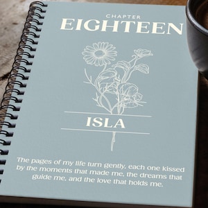 May include: A spiral-bound notebook with a light blue cover featuring the text "CHAPTER EIGHTEEN" and the name "ISLA". The cover also includes a floral illustration and a quote. A cup of coffee and a pen are next to the notebook.