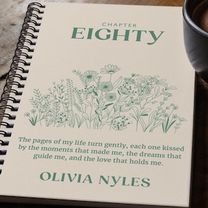Puede incluir: Una libreta espiral de color crema con un diseño floral verde y el texto "CHAPTER EIGHTY" en la parte superior. La libreta tiene una cita que dice "The pages of my life turn gently, each one kissed by the moments that made me, the dreams that guide me, and the love that holds me." y está firmada "OLIVIA NYLES".