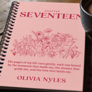 May include: A pink spiral bound notebook with the words "CHAPTER SEVENTEEN" in red lettering. The notebook has a floral design with a quote that reads "The pages of my life turn gently, each one kissed by the moments that made me, the dreams that guide me, and the love that holds me." The quote is followed by the name "OLIVIA NYLES" in red lettering.