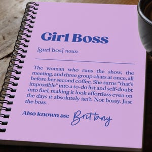 Może przedstawiać: Notatnik spiralny z jasnofioletową okładką z napisem Girl Boss, definicją w stylu słownikowym oraz imieniem Brittany zapisanym niebieskim tuszem. Notatnik znajduje się obok czarnego kubka z kawą i czarnego długopisu.