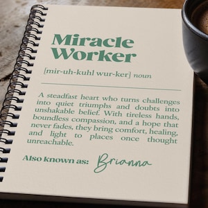 Puede incluir: Un cuaderno con espiral con las palabras "Miracle Worker" en verde. La definición de "Miracle Worker" está impresa debajo del título. Una taza de café y un bolígrafo también están sobre la superficie de madera.