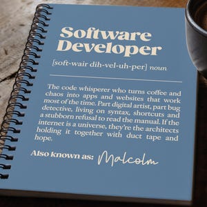 May include: A blue spiral-bound notebook featuring the text Software Developer, a phonetic pronunciation, a humorous definition, and the phrase Also known as: Malcolm in white lettering, positioned next to a black coffee mug and a black pen.