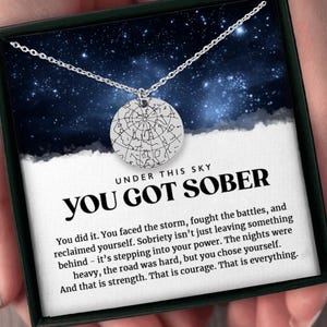 May include: A silver necklace with a circular pendant featuring a constellation design. The necklace is in a black box with a blue starry background and the text "UNDER THIS SKY YOU GOT SOBER". The text below the necklace reads "You did it. You faced the storm, fought the battles, and reclaimed yourself. Sobriety isn't just leaving something behind - it's stepping into your power. The nights were heavy, the road was hard, but you chose yourself. And that is strength. That is courage. That is everything."