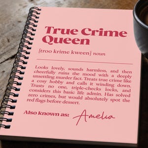 Puede incluir: Un cuaderno de espiral rosa con el texto "True Crime Queen" en granate. El cuaderno está abierto, revelando un texto que describe a un entusiasta del crimen real. Una taza de café negra con arte latte está cerca.